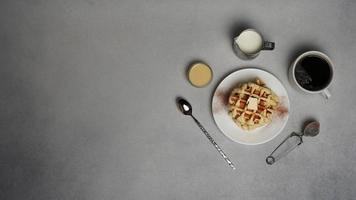 top visie van smakelijk wafels bord, karamel saus, koffie beker, melk, dessert lepel, zeef Aan een grijs beton achtergrond foto