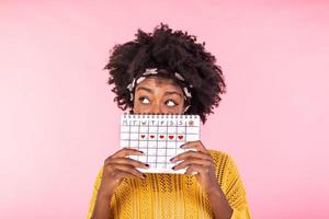 portret van een grappig jong Afrikaanse Amerikaans meisje in schuilplaats achter een menstruatie- periodes kalender en op zoek weg Bij kopiëren ruimte geïsoleerd over- roze achtergrond. vrouw periode kalender foto