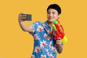 jong Aziatisch Mens Holding een water geweer en nemen een selfie met een smartphone Aan een geel achtergrond. Thais nieuw jaar dag of Thais songkran festival. foto