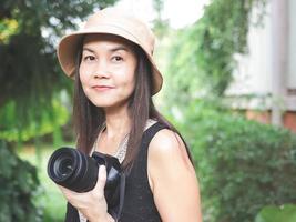 Aziatisch vrouw, vervelend hoed en zwart top mouwloos, staand in de tuin en Holding dslr camera, glimlachen gelukkig. foto
