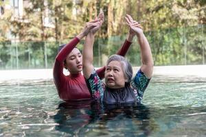 jong trainer helpen senior vrouw in aqua aerobics en werken uit in de zwembad. oud vrouw en volwassen Mens aan het doen aqua aerobics oefening in zwemmen zwembad, ouderen sport, en actief levensstijl concept. foto