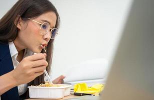 bezig en moe zakenvrouw aan het eten spaghetti voor lunch Bij de bureau kantoor en werken naar leveren financieel verklaringen naar een baas. overwerkt en ongezond voor klaar maaltijden, burn-out concept. foto