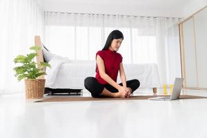 portret van gezonde jonge aziatische vrouw die yoga-oefeningen beoefent die in de slaapkamer zitten en thuis online op laptop leren. concept van lichaamsbeweging en ontspanning, technologie voor een nieuwe normale levensstijl foto