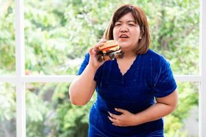 hongerig te zwaar vrouw Holding Hamburger Aan een houten bord, gedurende werk van huis, krijgen gewicht probleem. concept van eetbui aan het eten wanorde foto