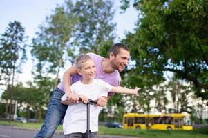 een vader ritten zijn zoon in een wit t-shirt Aan een scooter, de gelukkig kind verspreiding zijn handen foto