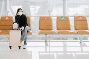 vrouw met gezichtsmasker zittend op sociale afstandsstoel met bagage foto