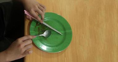 hand- Holding metaal ijzer bestek eetstokjes en lepel met afgewerkt aan het eten gebaar. leeg groen blad patroon bord Aan top van licht houten bureau tafel. geïsoleerd foto met gemakkelijk concept.