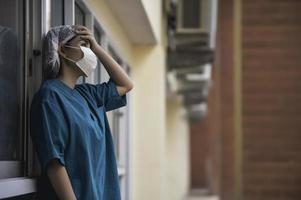 vermoeide depressieve vrouwelijke Aziatische scrubverpleegster draagt gezichtsmasker blauw uniform zit op de ziekenhuisvloer, jonge vrouwelijke arts gestrest van hard werken foto
