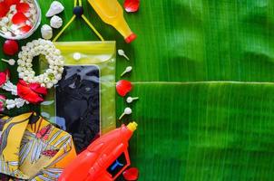 Thailand songkran festival achtergrond met smartphone in waterbestendig geval, water geweer, shirt, slinger en geparfumeerd water Aan banaan blad achtergrond. foto