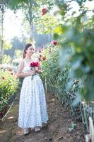 jong Aziatisch vrouw vervelend een wit jurk poses met een roos in roos tuin foto