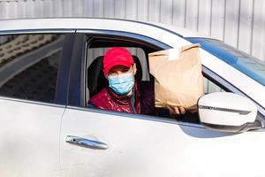 levering door auto. Mens in beschermend masker en medisch handschoenen Holding een papier doos. levering onderhoud onder quarantaine, ziekte uitbraak, coronavirus covid-19 pandemisch conditie. foto