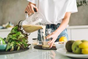 jong slank vrouw in wit t-shirt en blauw jeans Koken groen smoothie gezond voedsel in keuken Bij huis foto