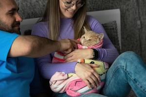 een jong paar Aan de bed knuffels hun kat verpakt in een baby deken en Toneelstukken met haar met een baby rammelaar. bewust weigering van een paar naar hebben kinderen, behandeling van huisdieren net zo kinderen. foto