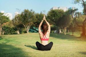 jong volwassen vrouw in sportkleding aan het doen yoga in de park buitenshuis, gezond vrouw zittend Aan gras en meditatie met lotus houding in ochtend. welzijn, fitheid, oefening en werk leven balans concepten foto