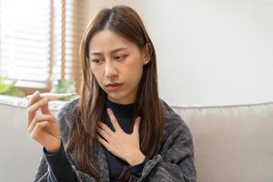 ziek, influenza Aziatisch jong vrouw, meisje hoofdpijn hebben een koorts, griep en controleren thermometer meten lichaam temperatuur, voelen ziekte zittend Aan sofa bed Bij huis. Gezondheid zorg persoon Aan virus, covid19. foto