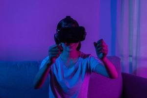 recreatie, Aziatisch jong vrouw vervelend vr koptelefoon of zichtbaar realiteit stofbril, bril of apparaatje, Speel video spel van simulator toekomstig, futuristisch terwijl draagbaar, zittend Aan bank, bank. foto