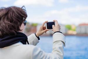 jong vrouw Aan vakantie gebruik makend van mobiel telefoon naar nemen afbeeldingen rivier- en stad foto