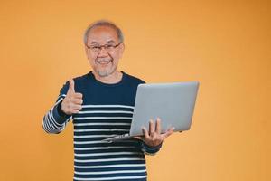 Aziatisch senior Mens gebruik makend van laptop computer voor werken na pensioen Aan de geel achtergrond. foto