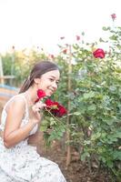 jong Aziatisch vrouw vervelend een wit jurk poses met een roos in roos tuin foto