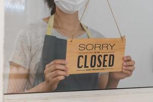 Aziatisch jong eigenaar detailhandel, koffie winkel vrouw draaien teken bord naar Gesloten met vervelend gezicht masker, bescherming naar pandemisch van coronavirus.sluiten winkel, restaurant of kroeg ten gevolge naar lockdown, quarantaine van covid. foto