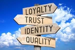 loyaliteit, vertrouwen, identiteit, kwaliteit - houten wegwijzer met vier pijlen, lucht met wolken in achtergrond foto
