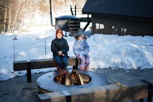 broer met zus zittend door kamp vreugdevuur Aan winter tegen klein huis. kinderen in platteland. foto