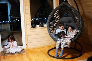 broer met zus lezing boeken in comfortabel hangende stoel in knus houten klein cabine huis. leven in platteland. foto