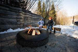 broer met zus zittend door kamp vreugdevuur Aan winter in Woud. kinderen in platteland. foto