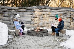 moeder met kinderen zittend en lezing boeken door kamp vreugdevuur Aan winter in Woud. kinderen in platteland. foto