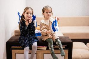 twee zussen zijn zittend Aan een bankstel Bij huis met Fins vlaggen Aan handen. Finland kinderen meisjes met vlag . foto