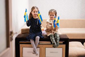 twee zussen zijn zittend Aan een bankstel Bij huis met Zweeds vlaggen Aan handen. Zweden kinderen meisjes met vlag . foto