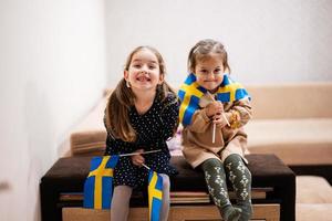 twee zussen zijn zittend Aan een bankstel Bij huis met Zweeds vlaggen Aan handen. Zweden kinderen meisjes met vlag . foto