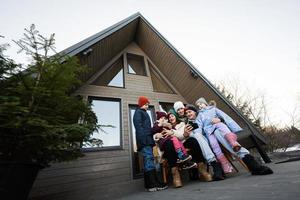 groot familie met vier kinderen zitten Aan terras uit rooster klein huis in de bergen op zoek Aan telefoons. foto