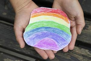 de handen van een klein kind zijn voorzichtig Holding een regenboog stuk van papier getrokken met gekleurde potloden. foto