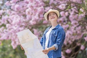 Aziatisch vrouw toerist Holding stad kaart terwijl wandelen in de park Bij kers bloesem boom gedurende voorjaar sakura bloem festival concept foto