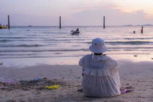 achterzijde visie van Aziatisch senior vrouw met baby speelgoed op zoek Bij zee terwijl zittend Aan strand. kopiëren ruimte, water sport, recreatie, pensioen, ongewijzigd, toneel, vakantie, genot, natuur. foto