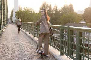 portret van jong bedrijf vrouw met een elektrisch scooter naar werk over- brug in modern stad achtergrond foto