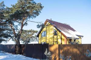 een geel knus huis in de sneeuw in winter in de dorp is omringd door pijnboom bomen. met sneeuw bedekt dak, verwarming en ventilatie pijpen, trapezium ramen foto