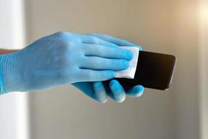 dichtbij omhoog is voor mannen is handen in medisch blauw rubber handschoenen naar wrijven een mobiel telefoon scherm met een servet. foto