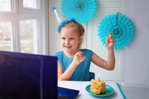 een weinig meisje zit achter een laptop, met een taart en kaars, dansen en vieren haar verjaardag over- de internetten. foto