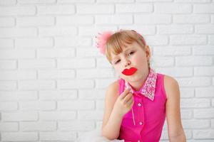 mooi weinig meisje, vervelend Aan roze jurk, poseren met rood papier lippen, met een kroon Aan zijn hoofd Aan wit achtergrond, taille omhoog. foto