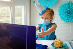 een gelukkig weinig meisje, vervelend een medisch masker in voorkant van haar laptop viert haar verjaardag via de internet in quarantaine tijd, zelfisolatie, online verjaardag. foto