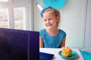 een weinig meisje zit achter een laptop, met een taart en kaars en vieren haar verjaardag over- de internetten. foto