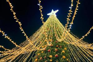 Kerstmis boom met geel slingers, decoratief bollen en groot wit ster topper Bij nacht blauw lucht foto