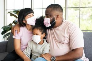 gemengd ras familie zittend Aan de sofa en vervelend medisch gezicht maskers voor tegen coronavirus wereld pandemisch en blijven in quarantaine geplaatst samen Bij huis sociaal afstand nemen nieuw normaal levensstijl foto