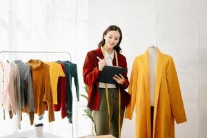 Aziatisch kleermaker vrouw werken Aan kleren in maatwerk atelier. mooi jong vrouw mode ontwerper glimlach en na succes in studio foto