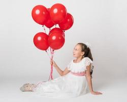 mooi meisje gekleed in lang wit jurk Holding veel van rood ballonnen in hand, op zoek omhoog Bij ballen foto