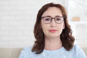 een midden- oud vrouw vervelend bril looks vriendelijk Bij de camera terwijl zittend Bij huis Aan de bankstel foto