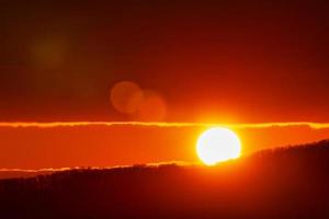 mooi zonsopkomst in bergen, rood zon schijf stijgt van tops van berg bereik. natuurlijk lens gloed in lucht foto