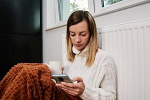 bezorgd vrouw zitten in de buurt verwarming radiator onder deken en gebruik makend van smartphone foto